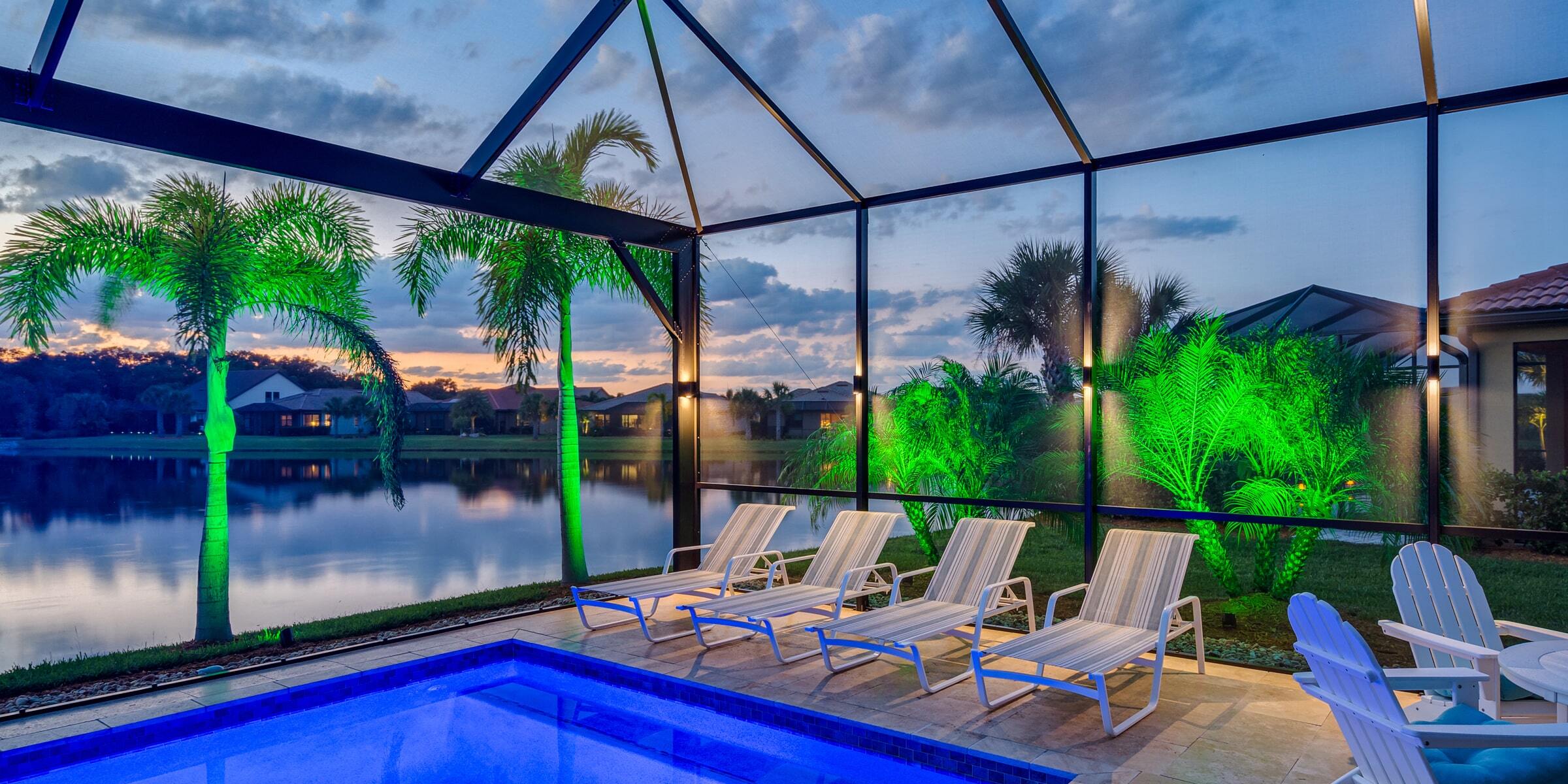 Pool cage at dusk with color-changing lighting on palms overlooking the water