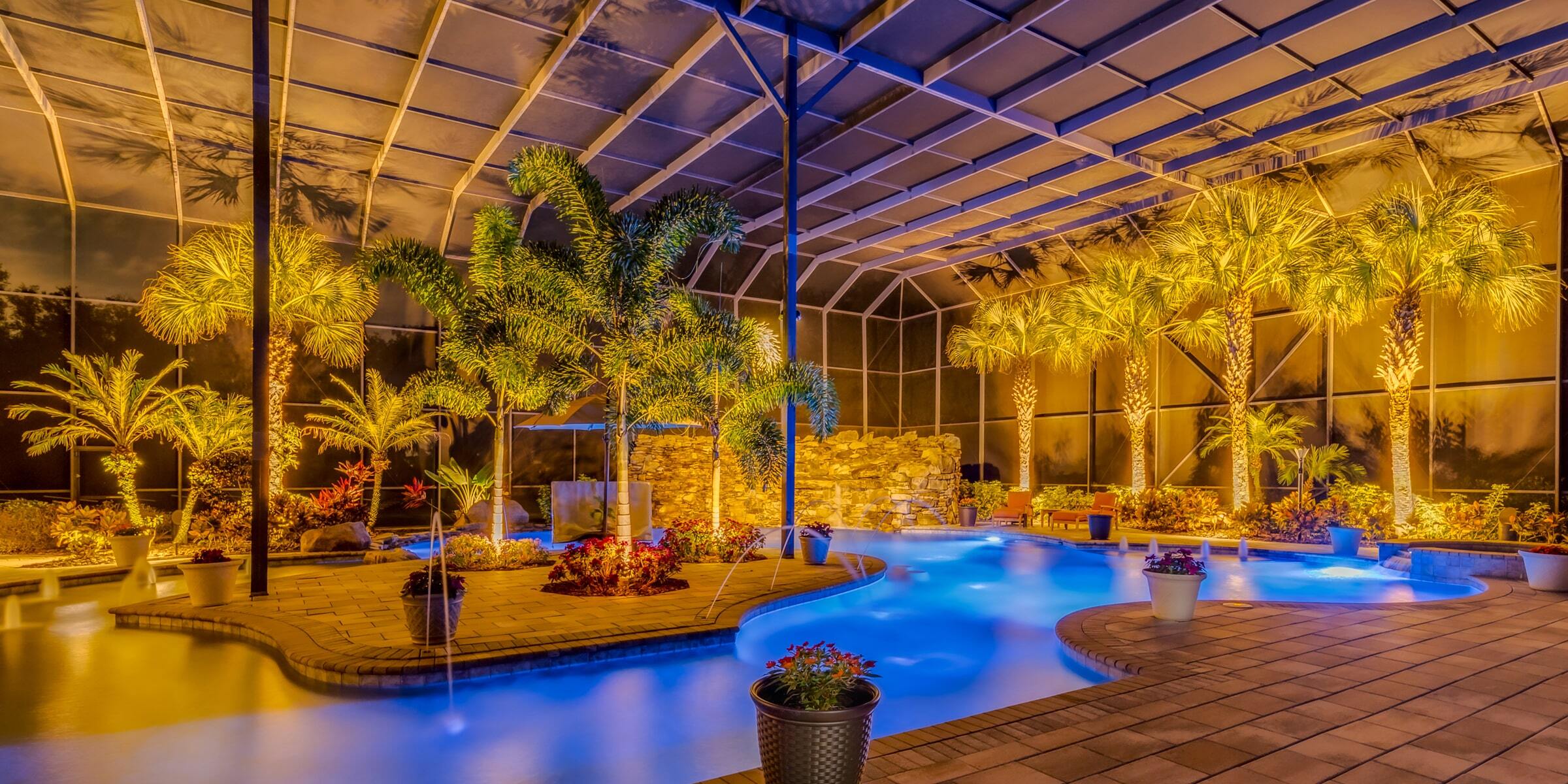 Screened pool enclosure with palms and tropical planting washed in warm light at night