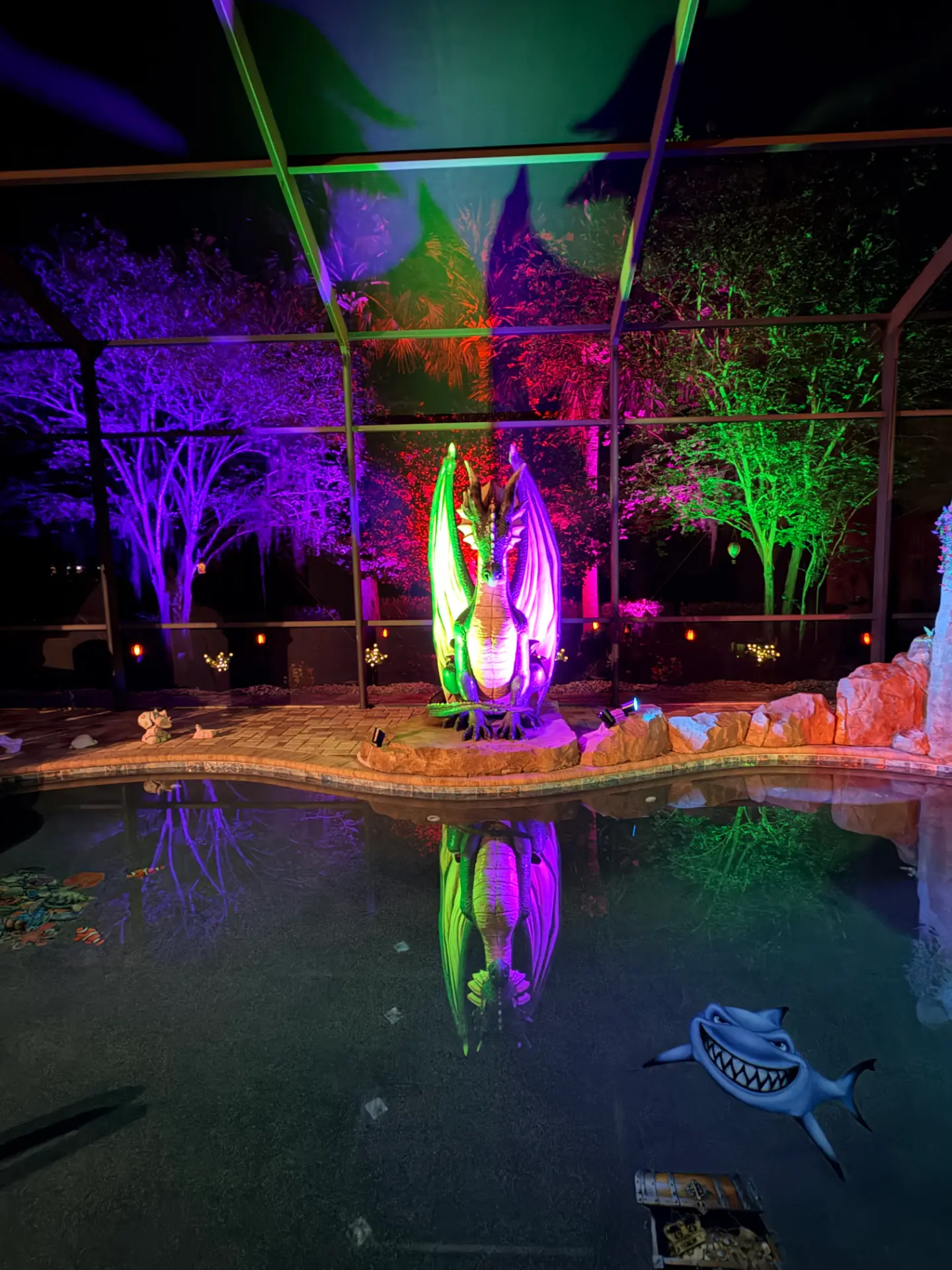 Pool enclosure and trees lit in full color above a pool at night