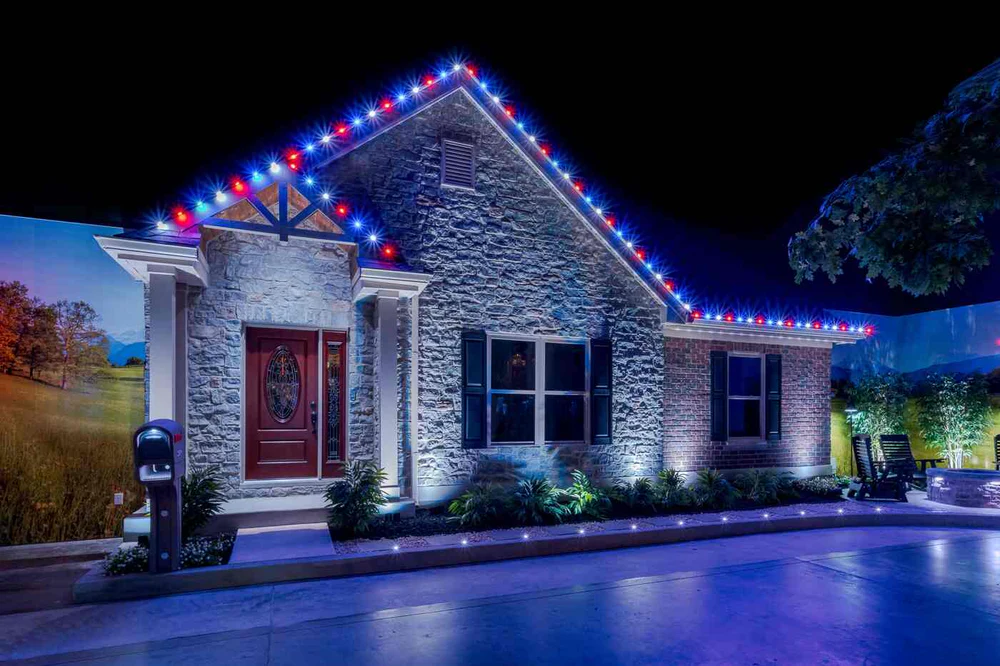 Red, white, and blue holiday lighting along the roofline of a stone home at night